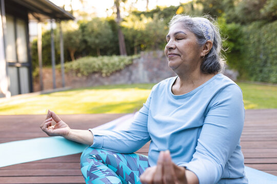 Senior woman practicing meditation on yoga mat on wooden deck in backyard garden, wearing headband