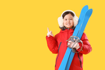 Little Asian girl with skis pointing at something on yellow background