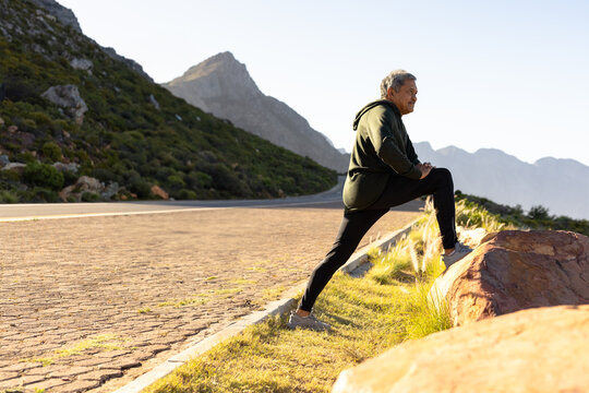 Senior multiracial man leaning on boulder and stretching leg on mountain road shoulder, copy space