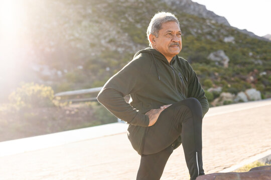 Senior man stretching right leg on mountain road, wearing hoodie using rock for support, copy space