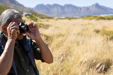 Senior man holding 35mm camera and framing shot in coastal meadow with golden grass, copy space