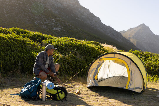 Senior man kneeling in mountain campsite, tightening dome tent guy line near backpacks, copy space