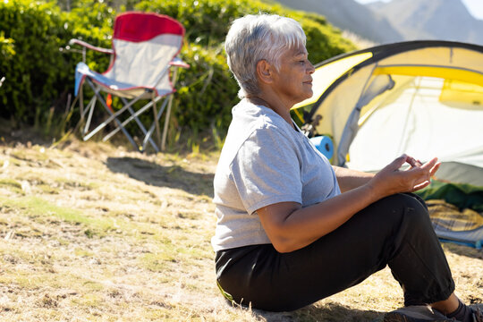 Senior woman sitting cross-legged at campsite, meditating with yellow-green tent and red chair