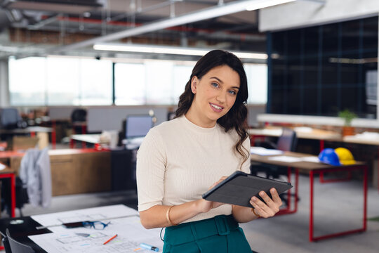 Woman holding tablet above desk with blueprints and hard hats in open-plan office, copy space