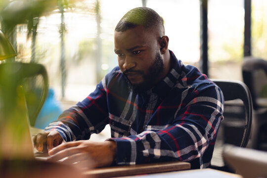 African American man typing on laptop at wooden desk in office, with potted plant in view