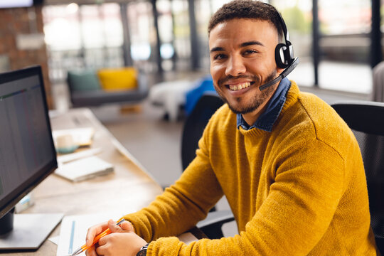 Man working at desk in office wearing headset using computer monitor, writing on notepad