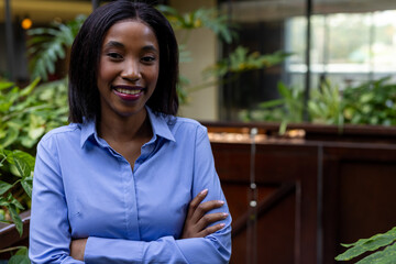 Young adult African American woman standing with crossed arms in office atrium, among potted plants