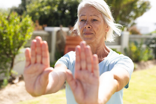 Senior female performing forward arm stretch in sunny backyard garden, practicing gentle exercise