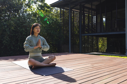 Asian woman meditating in prayer pose on wooden backyard deck, with yoga mat, copy space
