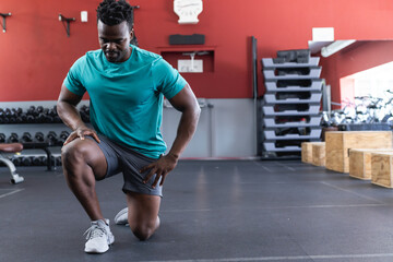 African American man kneeling in gym stretching, with dumbbell rack and step platforms, copy space