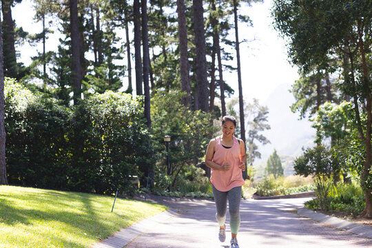 Mid adult Asian woman jogging on paved park path lined with pine trees, wearing running shoes
