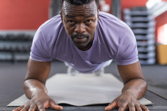 African American man holding forearm plank on gray mat in gym, with dumbbell rack, step platforms