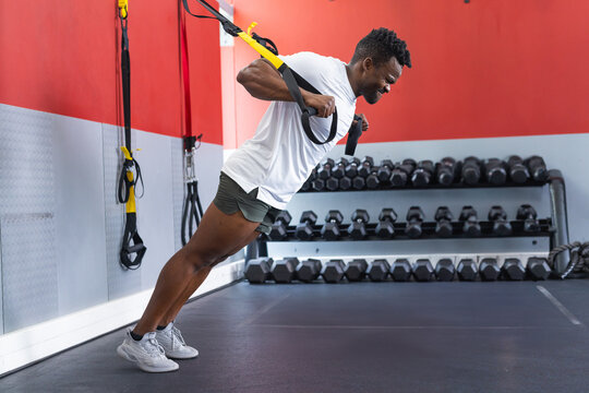African American man performing TRX row in gym corner with yellow black straps and dumbbell rack