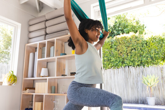 Teenage girl practicing aerial yoga in home studio near windows, gripping teal silk hammock