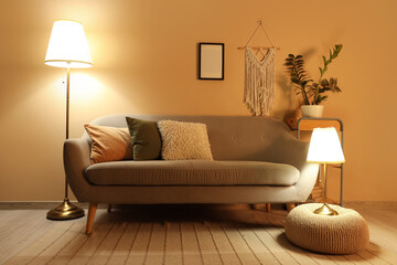 Interior of stylish living room with grey sofa, pouf and glowing lamps at evening