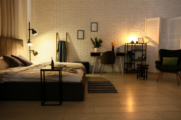 Stylish interior of bedroom with soft grey bed, table and glowing floor lamp at night