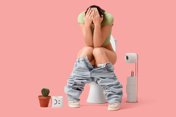 Suffering young woman sitting on toilet bowl against pink background. Cystitis concept