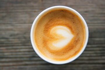 Top down view of a paper coffee  cup, placed on a rustic wooden table, filled with a creamy latte featuring heart shaped latte art