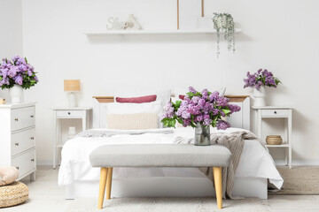 Interior of light bedroom with soft bench and lilac flowers in vases