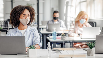 Fototapeta premium Work on project in corporate company after lockdown. African american woman in protective mask creating idea at workplace with smartphone and protective glass in office interior, free space