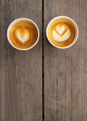 Top down view of two paper coffee cups, placed on a rustic wooden table, with a creamy heart and flower shaped latte art