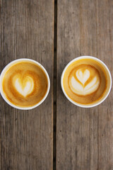 Top down view of two paper coffee cups, placed on a rustic wooden table, with a creamy heart and flower shaped latte art