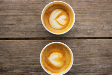 Top down view of two paper coffee cups, placed on a rustic wooden table, with a creamy heart and flower shaped latte art