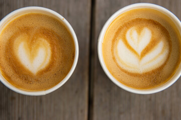 Top down closeup view of two paper coffee cups, placed on a rustic wooden table, with a creamy heart and flower shaped latte art  
