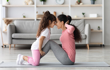 Loving middle eastern mother and daughter in sportswear working on abs together, exciting little girl holding her mom legs while she doing crunches, touching each other with foreheads, home interior