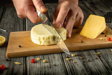 A person uses a knife to skillfully cut a round piece of cheese on a wooden cutting board. Nearby,...