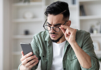 Eyesight Problems Concept. Young Arab Man In Eyeglasses Looking At Smartphone Screen And Frowning,...