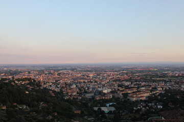 Obraz premium Panoramic view from Città Alta in Bergamo: the city stretches out in the warm sunset light, with rooftops, historic buildings, and flatlands fading into the horizon under a clear sky.