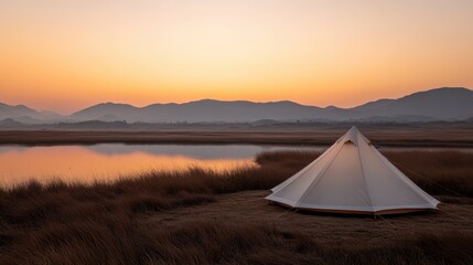 A tranquil scene featuring a tent by a calm body of water at sunrise, surrounded by mountains and tall grasses, Ideal for outdoor, camping, or nature-related projects,