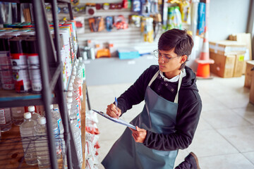 Focused male employee writing notes on clipboard while working in hardware store and controlling...