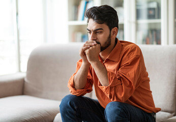 Fototapeta premium Permacrisis, life problems, financial hangout concept. Pensive young middle eastern man sitting on sofa at home, leaning on his hands, experiencing difficulties in life, looking at copy space