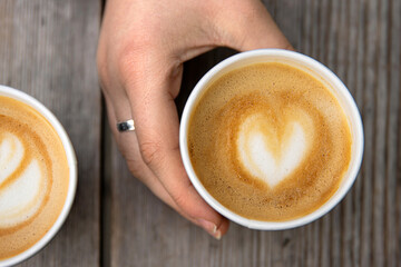 Closeup of a hand holding a paper coffee cup with heart-shaped latte art beside another cup on a rustic wooden table.