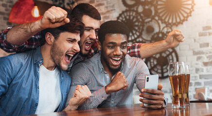 Diverse football fans watching football on smartphone and celebrating victory score in pub