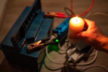 Hand with candle near battery powering devices in blackout. Hand holding candle near car battery with inverter and phones charging, symbolizing emergency lighting and power backup during blackout