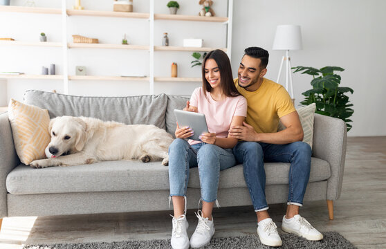 Beautiful young diverse couple sitting on couch with their cute dog, using tablet pc, browsing web at home, copy space. Millennial multiracial spouses with touch pad surfing internet indoors