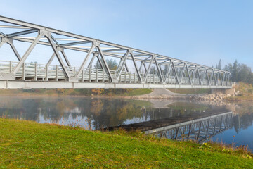 A pedestrian deck with metal railings spans a tranquil, narrow river. The bridge adjoins two riverbanks. Steam rises from the water, reflecting the structure in the water. 