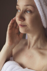 Fototapeta premium A woman with a towel on her head, after a bath, looks away with a gentle expression. A woman wrapped in a white towel after a bath. Spa day relaxing.