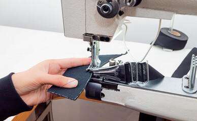 Close-up of a person sewing heavy fabric on an industrial sewing machine. The needle, presser foot, and hands are shown working on a piece of textile in a workshop. Shoe sewing machine, cylindrical.