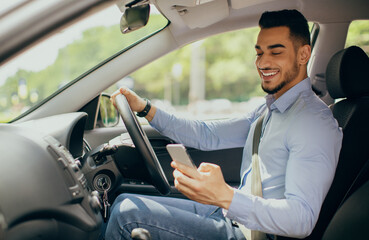 Bad driving habits, unsafe driving, driving and gadgets concept. Cheerful arab man entrepreneur going to office by auto, using cellphone while driving car, chatting with someone, side view, copy space
