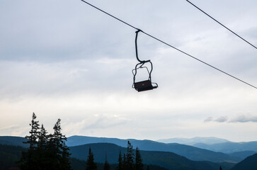 Single ski chairlift hangs suspended against a cloudy sky, with forested mountains in the background creating a peaceful and isolated atmosphere