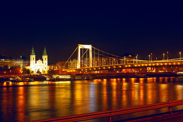 Obraz premium Elisabeth Bridge in Budapest stretches over Danube River, suspension cables glowing under night sky. City twinkling skyline, with historic buildings and distant cathedral creates stunning backdrop