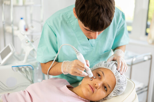 Young male cosmetologist performs hardware facial microcurrent procedure to young female patient