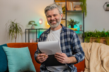 Smiling bearded mature man in casual clothes with documents analyzing information reading business...