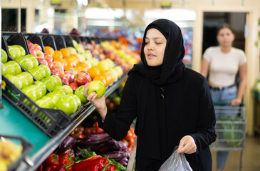In supermarket,middle-aged woman in traditional Muslim hijab takes and puts ripe apples in small bag