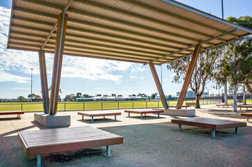 Modern outdoor pavilion structure offers shade over timber slat benches in a public sports reserve in Melbourne, Australia. Public community recreational infrastructure.
