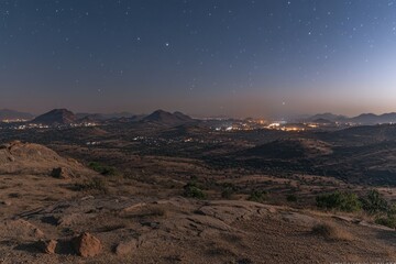 Nighttime panorama of a dry, hilly landscape, city lights visible in the distance, under a starry sky